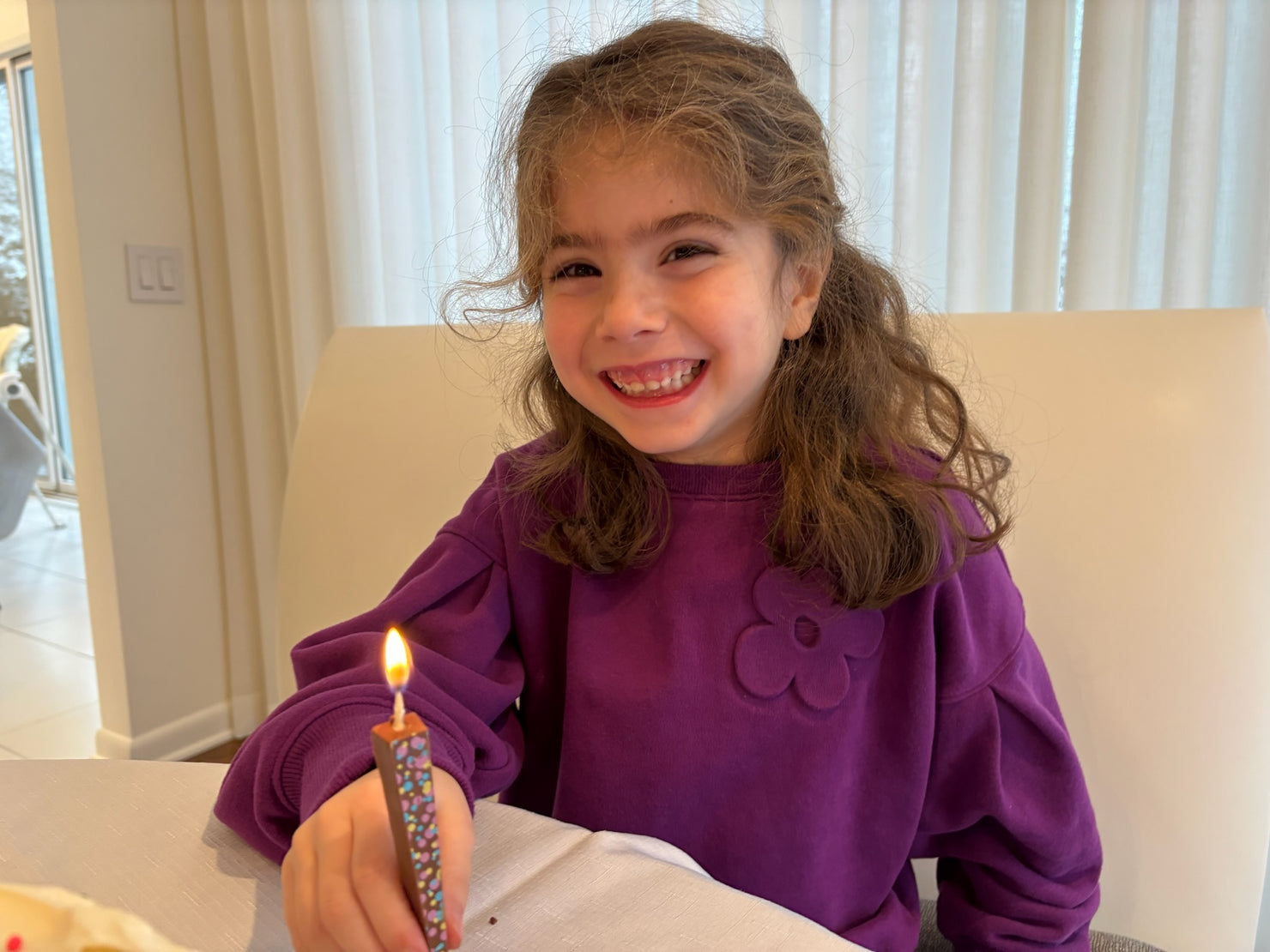 A young girl with brown hair smiles while holding a lit milk chocolate birthday candle with tri-color dots. She is wearing a purple sweatshirt and sitting indoors in front of a white chair and window blinds.