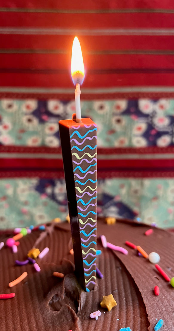 A single tall dark chocolate candle with a tri-color wave pattern, lit and standing in chocolate cake frosting with colorful sprinkles, against a patterned red and floral background.